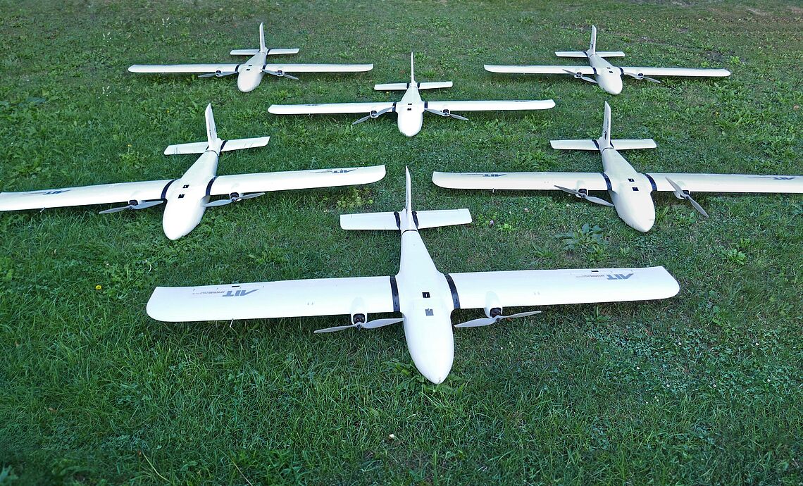 Mehrere Modellflieger mit Sensorik und AIT Logo ausgestatt liegen auf der Wiese vor dem AIT Fluglabor