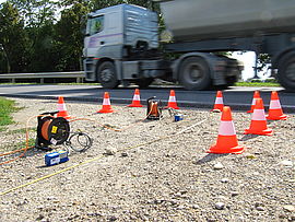 A road where traffic cones stand, which are arranged in the shape of a rectangle with the front side open. In the background a truck drives by. In the rectangle there are measuring devices. 