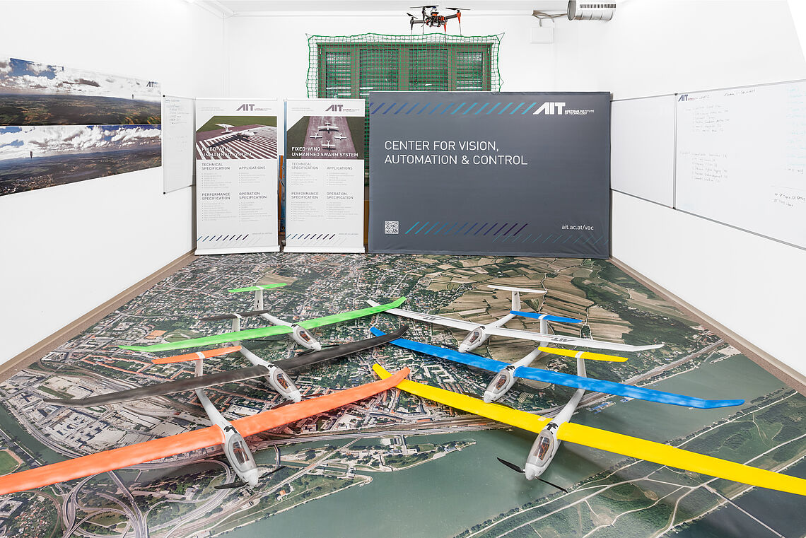 Verschiedenfärbige Modellflieger liegen auf dem Boden auf einem großen Luftbild im AIT Fluglabor