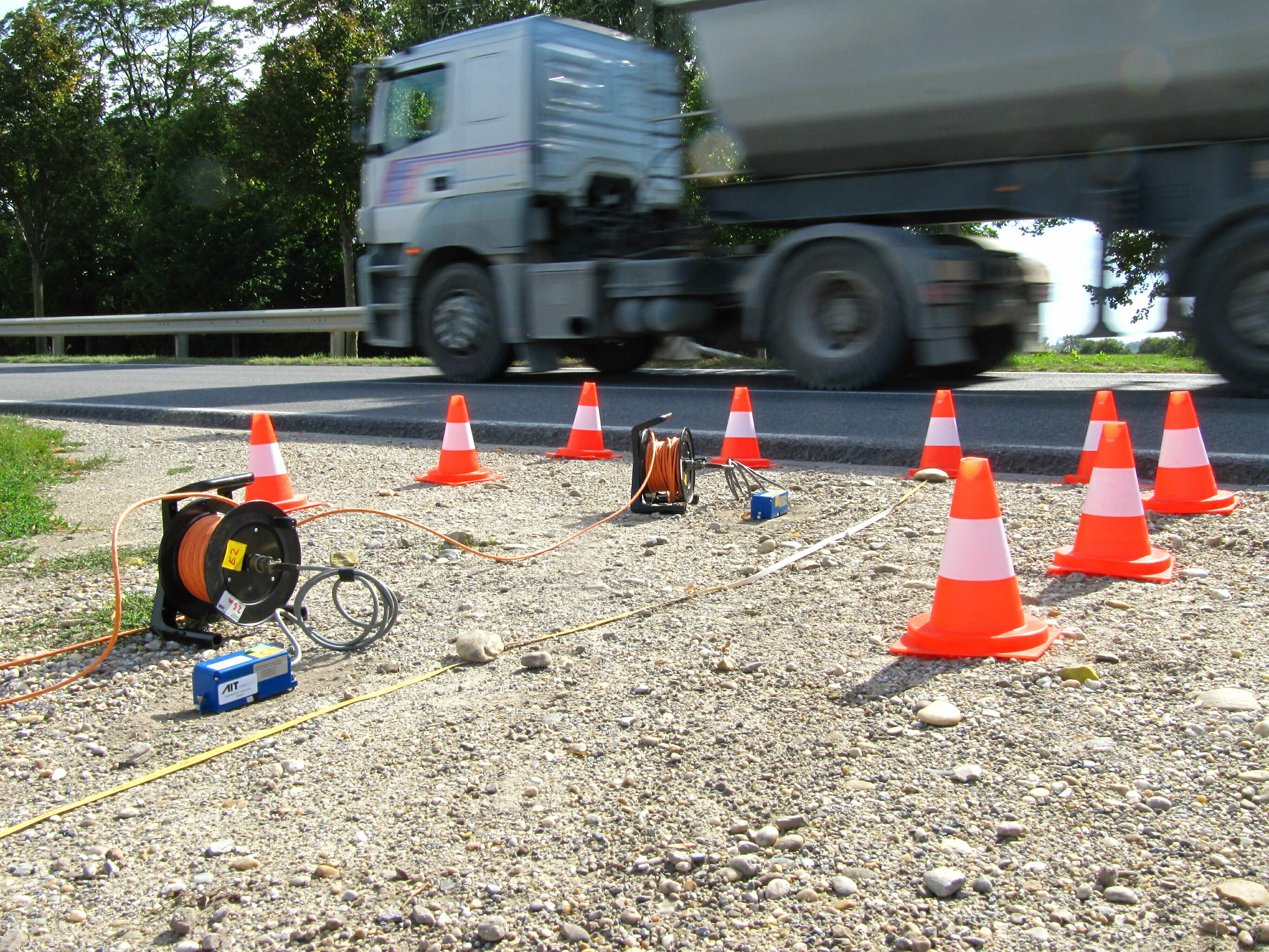 Eine Straße an der Verkehrshütchen stehen, die in Form eines Rechtecks aufgestellt sind beid er die vordere Seite offen ist. im Hintergrund fährt ein LKW vorbei. Im Rechteck sind Messaperaturen angebrach. 