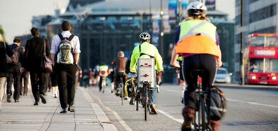 ein Gehweg auf dem viele Fußgänger unterwegs sind, daneben ist ein Fahrradstreifen auf dem viele Radfahrer fahren. Daneben befindet sie eine Straße auf der Autos zu sehen sind.